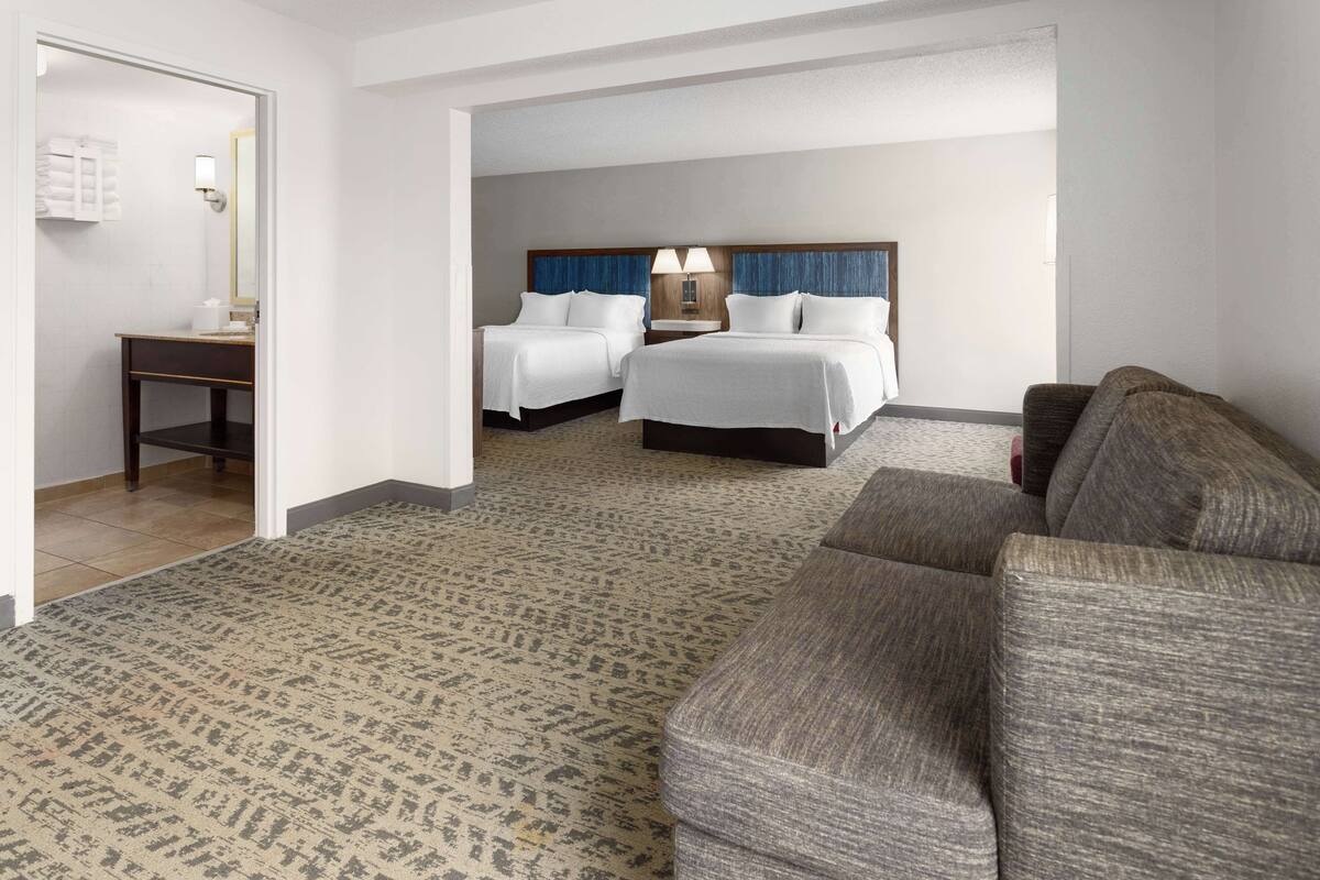 A hotel room with two beds, a sofa, a small table, and a bathroom visible through an open door.