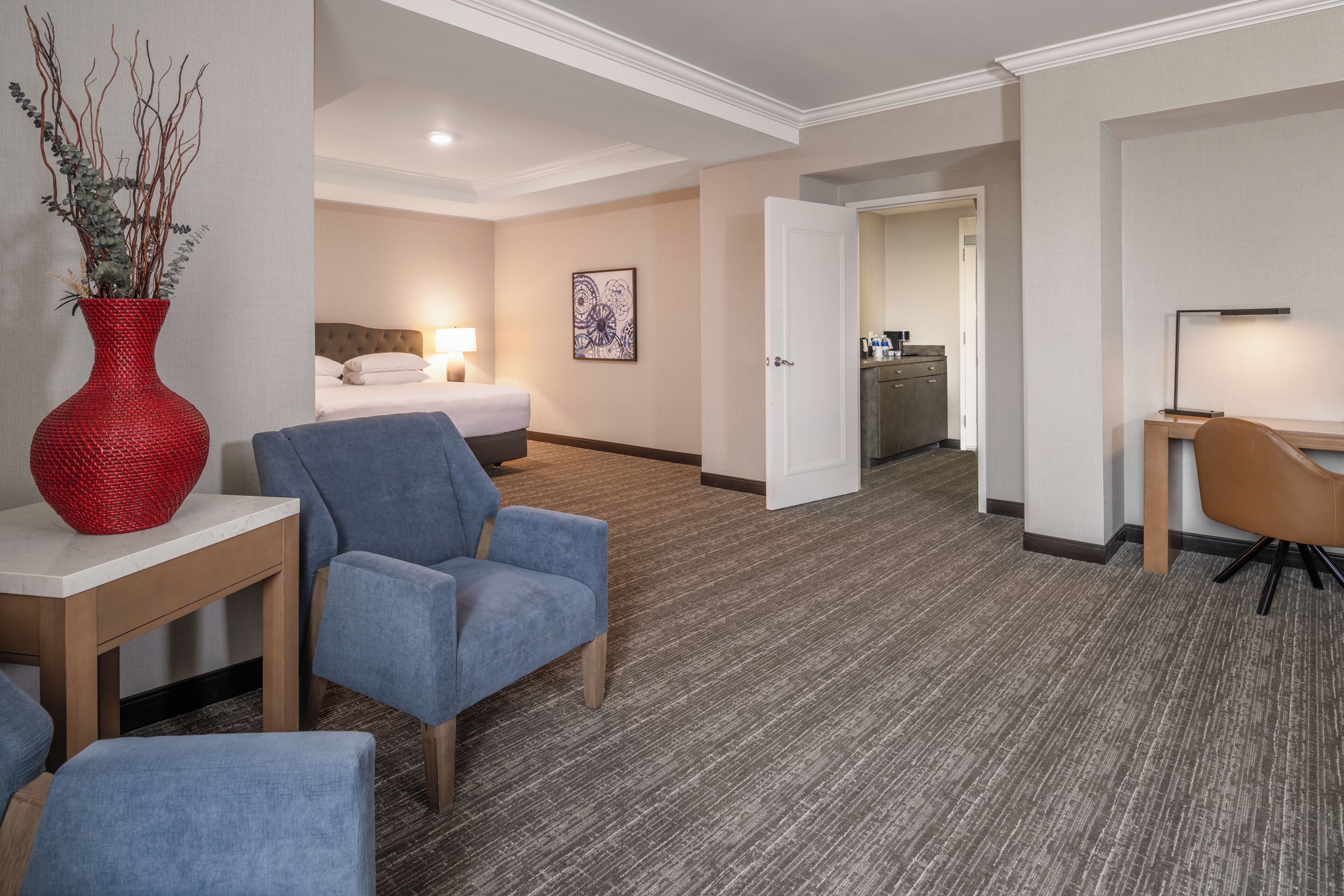 A hotel room with a bed, a desk, a chair, a red vase, and a painting on the wall.