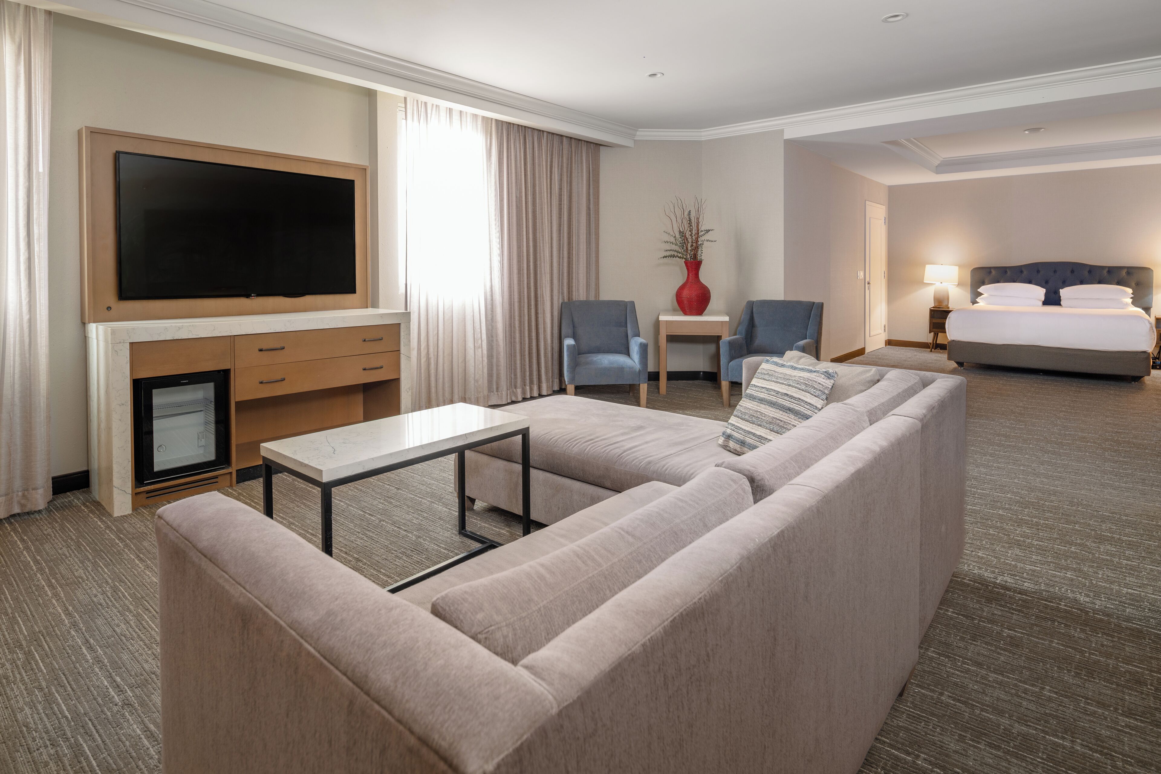 A hotel room with a large flat-screen TV, a sofa, two armchairs, a small table, a bed, and a nightstand with a lamp.