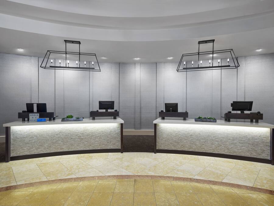 Zona de recepción con dos mostradores, cada uno con una computadora y una planta pequeña. Dos lámparas colgantes iluminan los mostradores, y las paredes están cubiertas de azulejos.