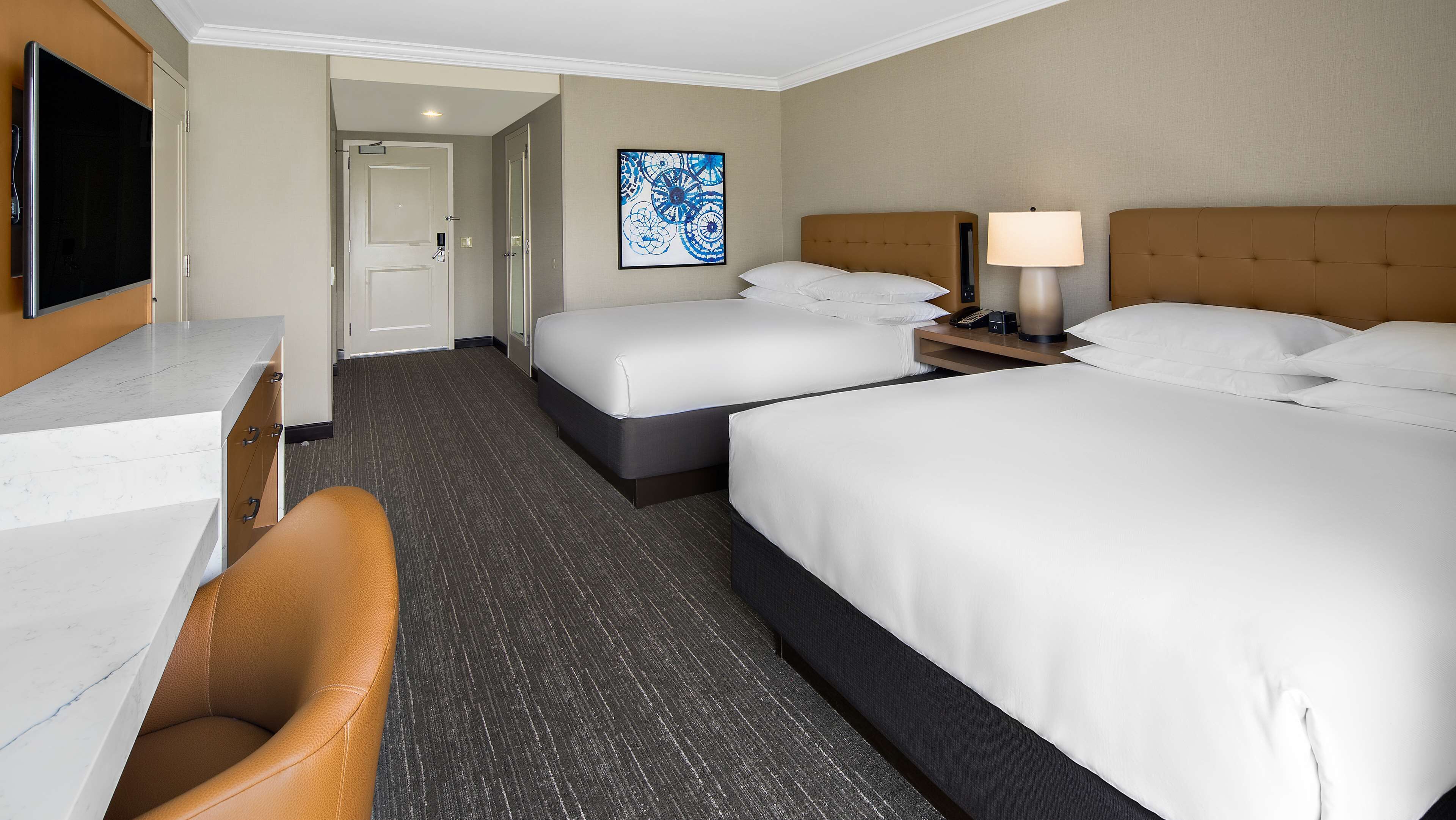 A hotel room with two beds, a desk, a chair, a TV, and a painting on the wall.