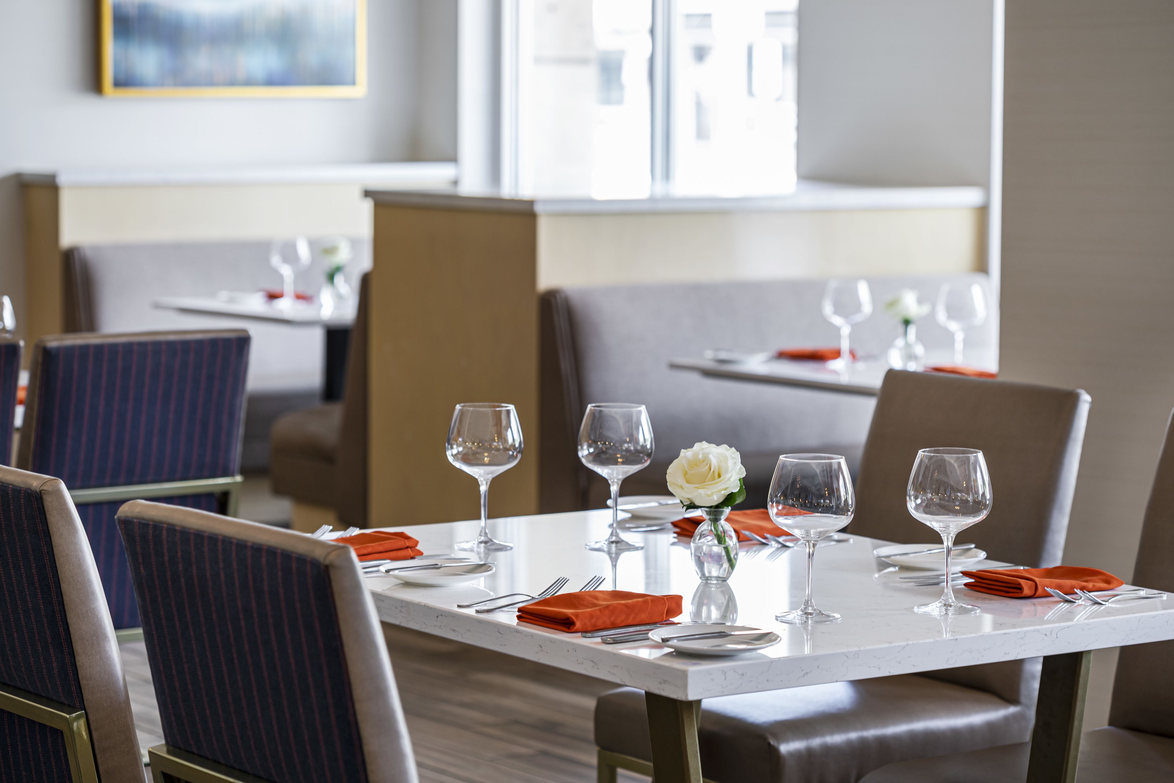 A restaurant setting with tables set for dining, featuring chairs, wine glasses, and a vase with a flower.