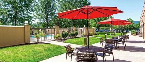 An outdoor patio area with tables and chairs, red umbrellas, and a swimming pool in the background.