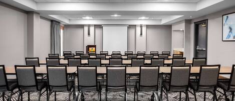 Un salón de conferencias con varias mesas y sillas, una pantalla de proyector y piso alfombrado.