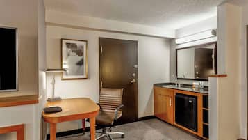 A hotel room with a wooden desk, a chair, a small refrigerator, a sink, and a mirror.