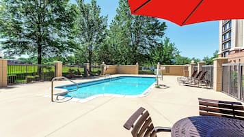 A swimming pool with lounge chairs and a red umbrella, surrounded by trees and a fence.