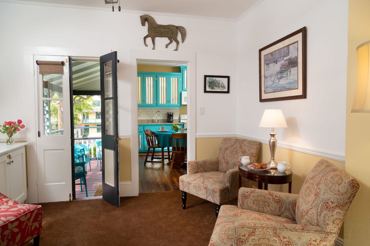 A living room with two patterned armchairs, a coffee table, and a door leading to a kitchen with teal cabinets.