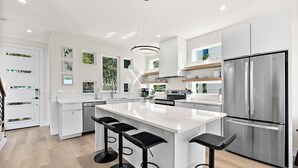 Une cuisine moderne dotée d’un grand îlot central, de tabourets de bar, d’appareils électroménagers en acier inoxydable et d’une palette de couleurs épurée, dans des tons blancs.