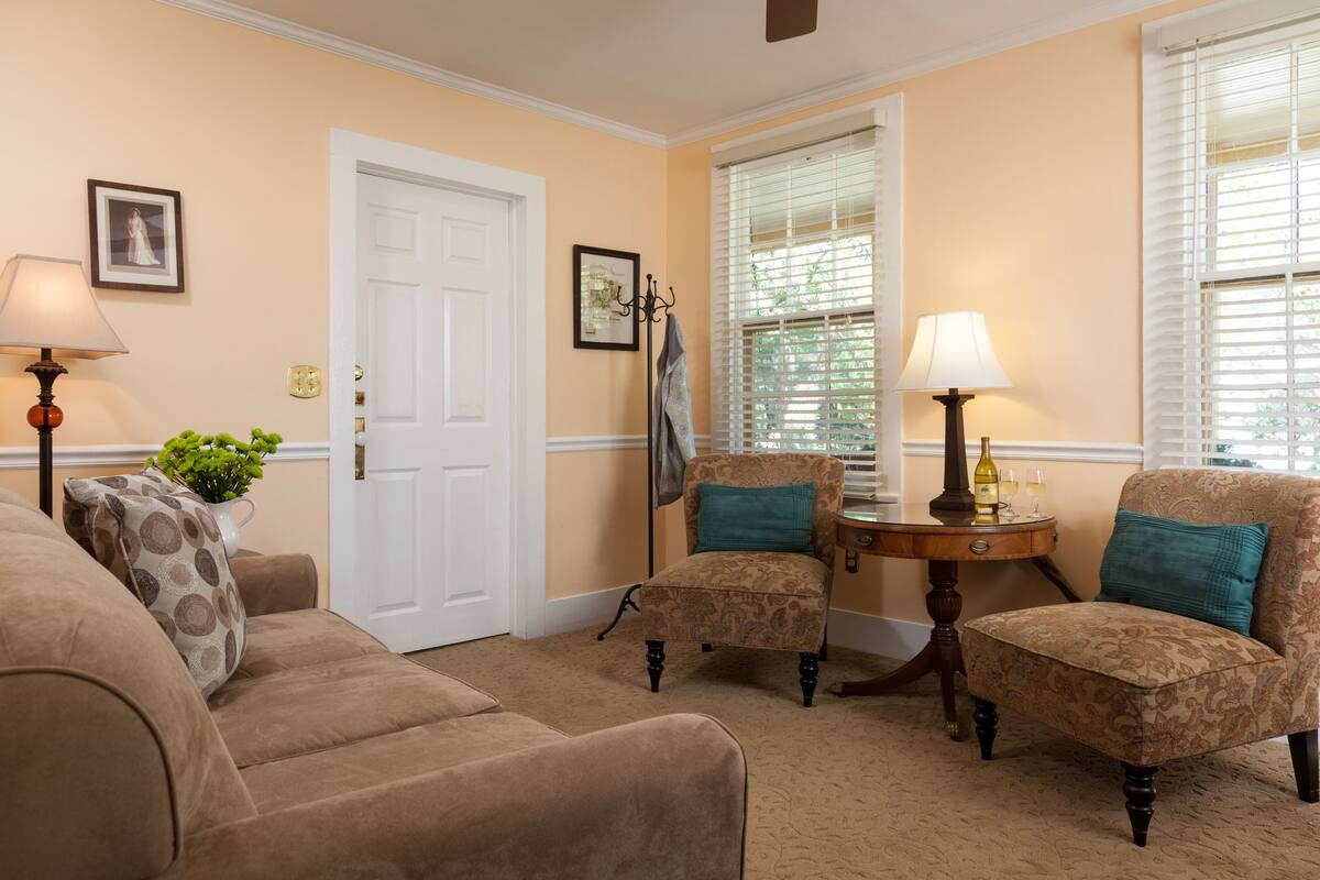 A living room with a beige carpet, two patterned armchairs, a sofa, a white door, and a small table with a lamp and a bottle.