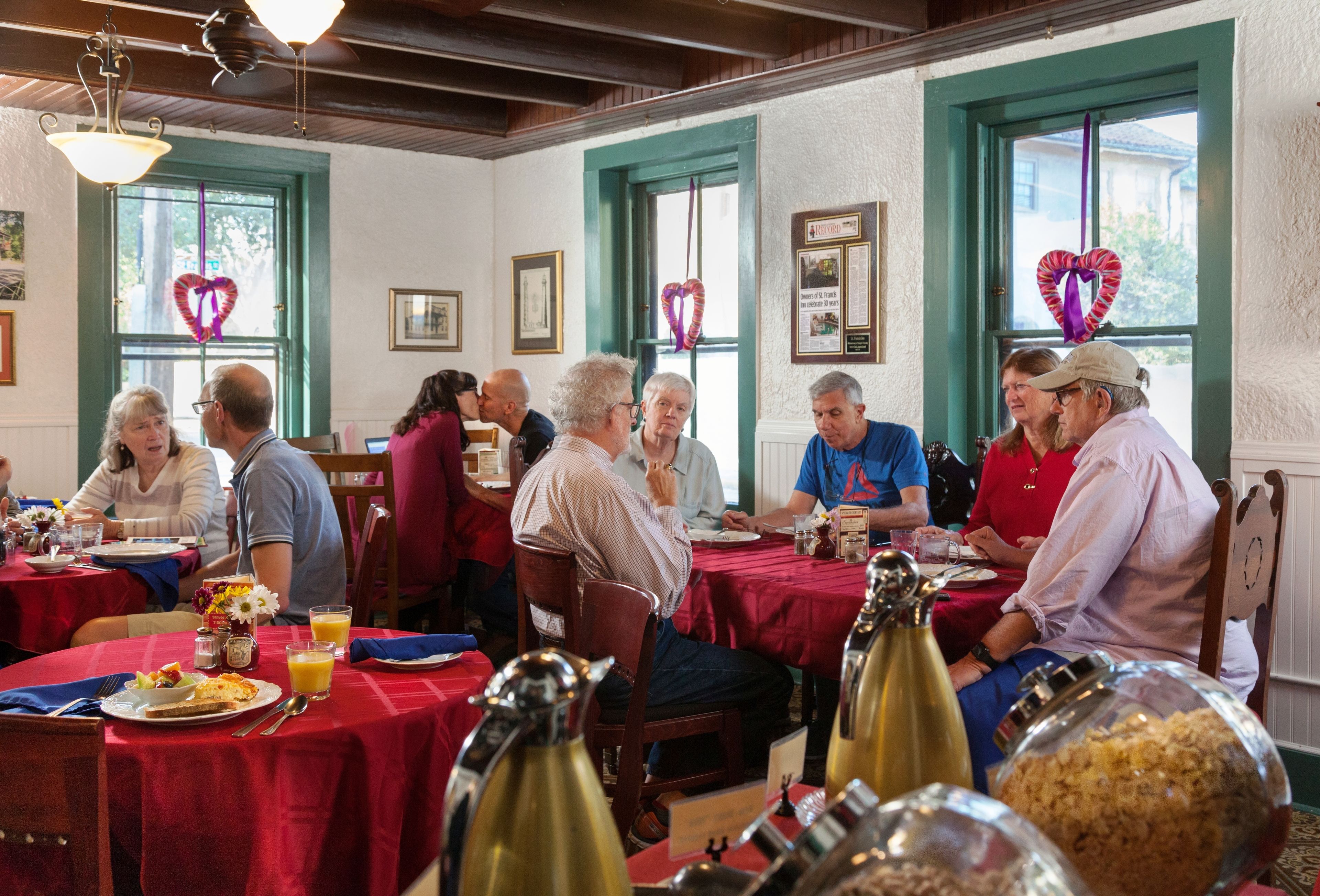 A dining area with round tables covered in red tablecloths, wooden chairs, and various food and drink items.