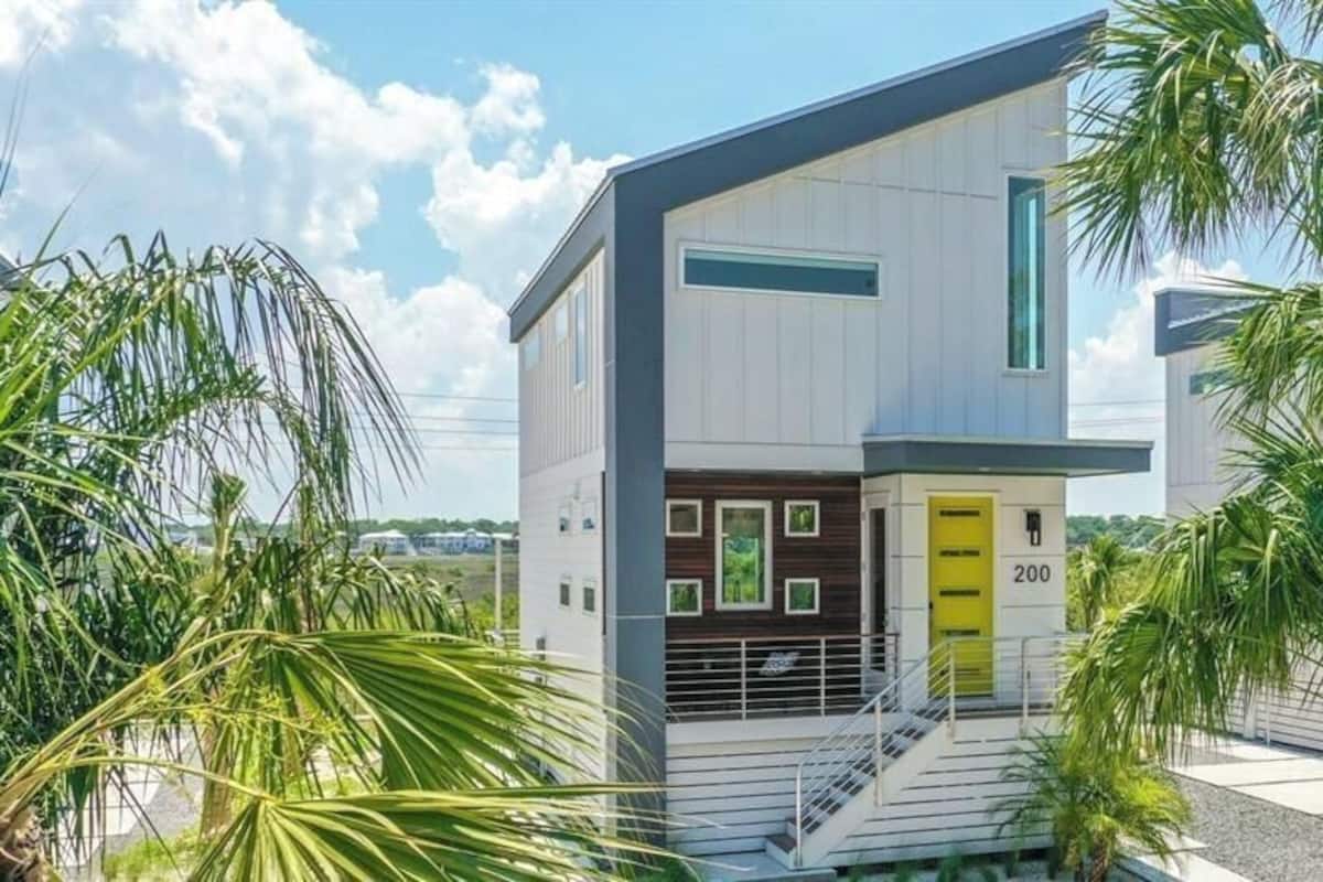 A modern two-story house with a yellow door, a staircase leading to the entrance, and a view of palm trees.