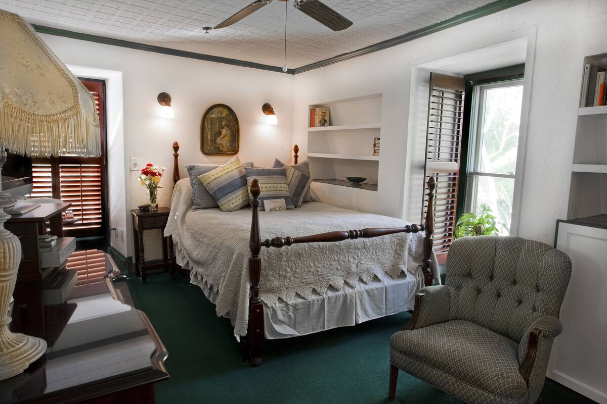 A bedroom with a four-poster bed, a wooden chair, a desk with a lamp, and a bookshelf.