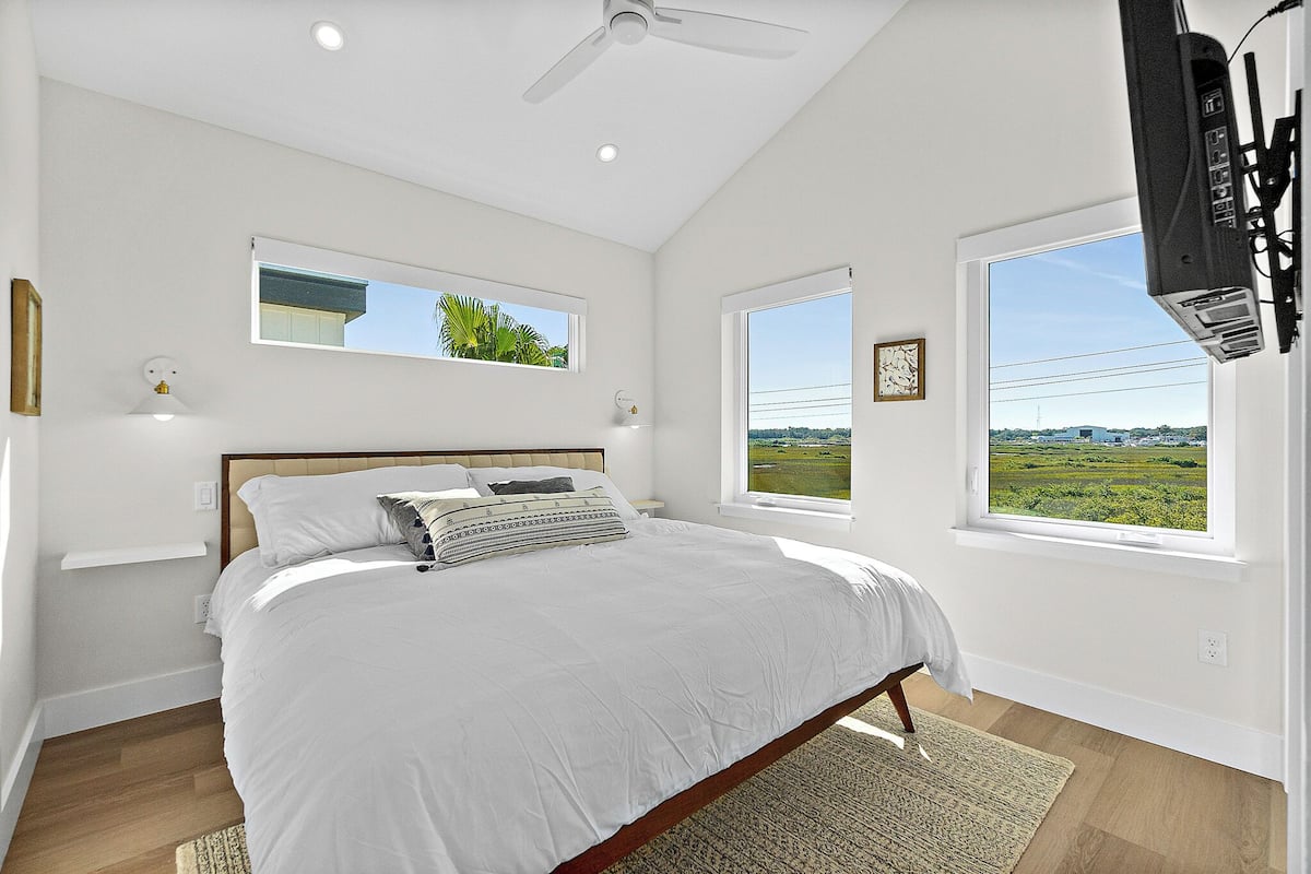 A bedroom with a large bed, two windows with views, a ceiling fan, and a mounted TV.
