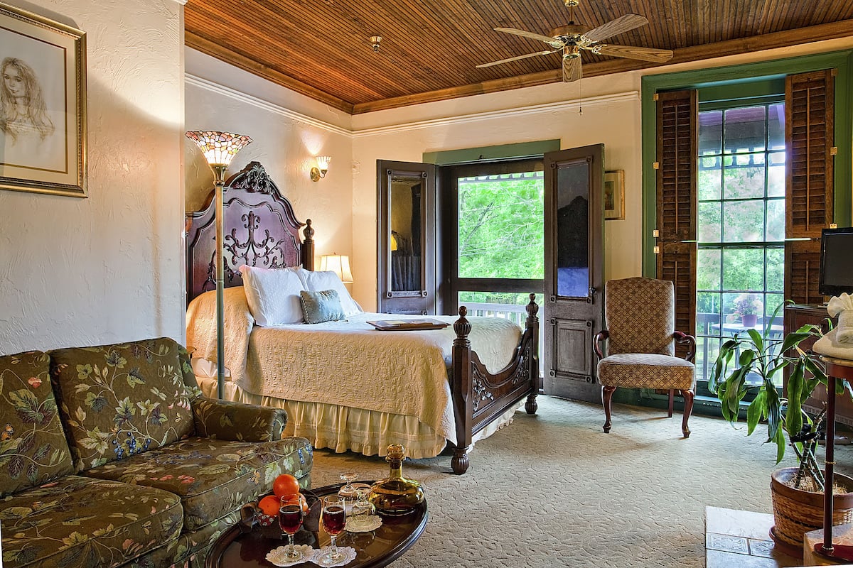 A bedroom with a bed, a chair, a sofa, a window with shutters, and a ceiling fan.