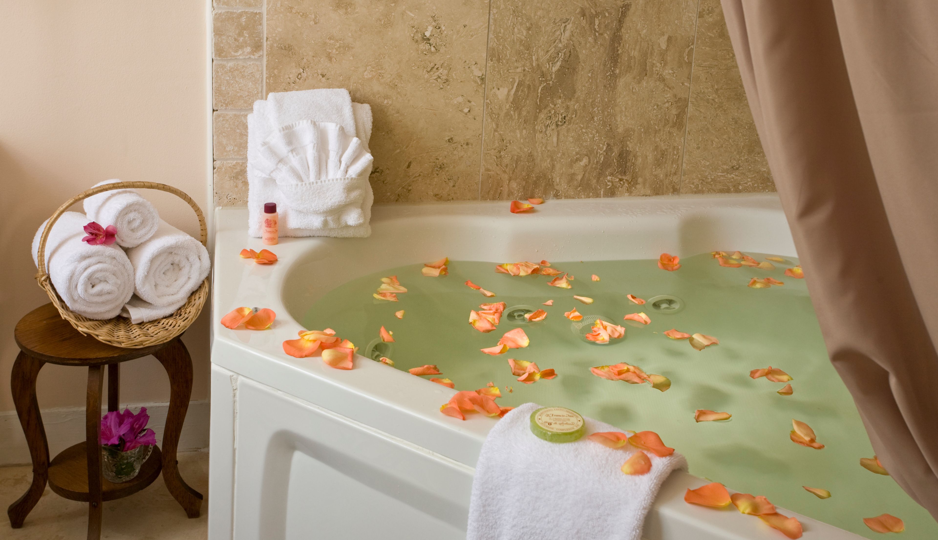 A bathroom with a bathtub filled with rose petals, a basket of rolled towels, and a small table with a vase of flowers.