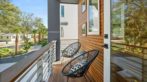 Un balcon doté de deux fauteuils en osier et d’une balustrade en bois, donnant sur un quartier résidentiel avec des palmiers et des maisons.