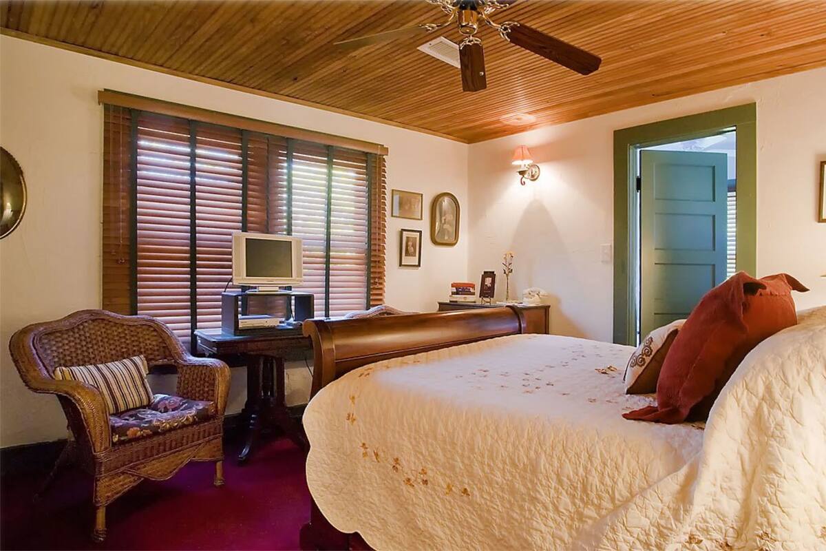 A bedroom with a bed, a chair, a desk with a computer, and a ceiling fan.