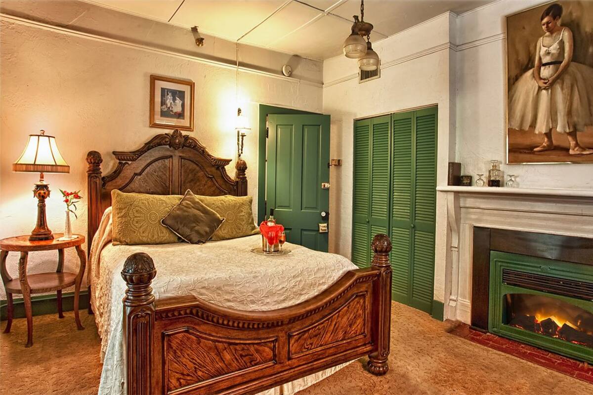 A bedroom with a wooden four-poster bed, green shutters, a fireplace, and a framed portrait on the wall.