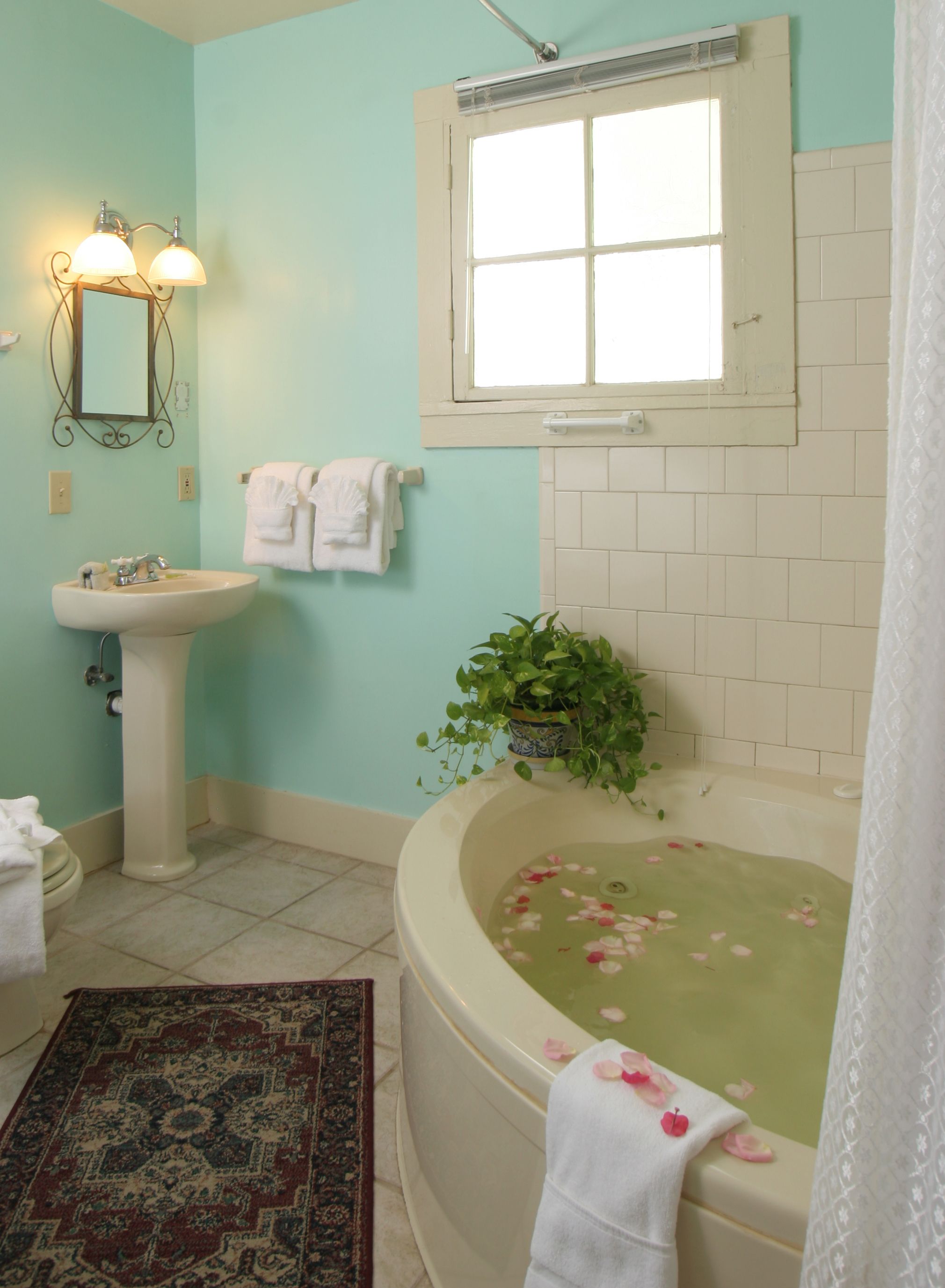 Un baño con lavamanos, espejo, ventana y bañera con pétalos de rosa, además de una planta en maceta.