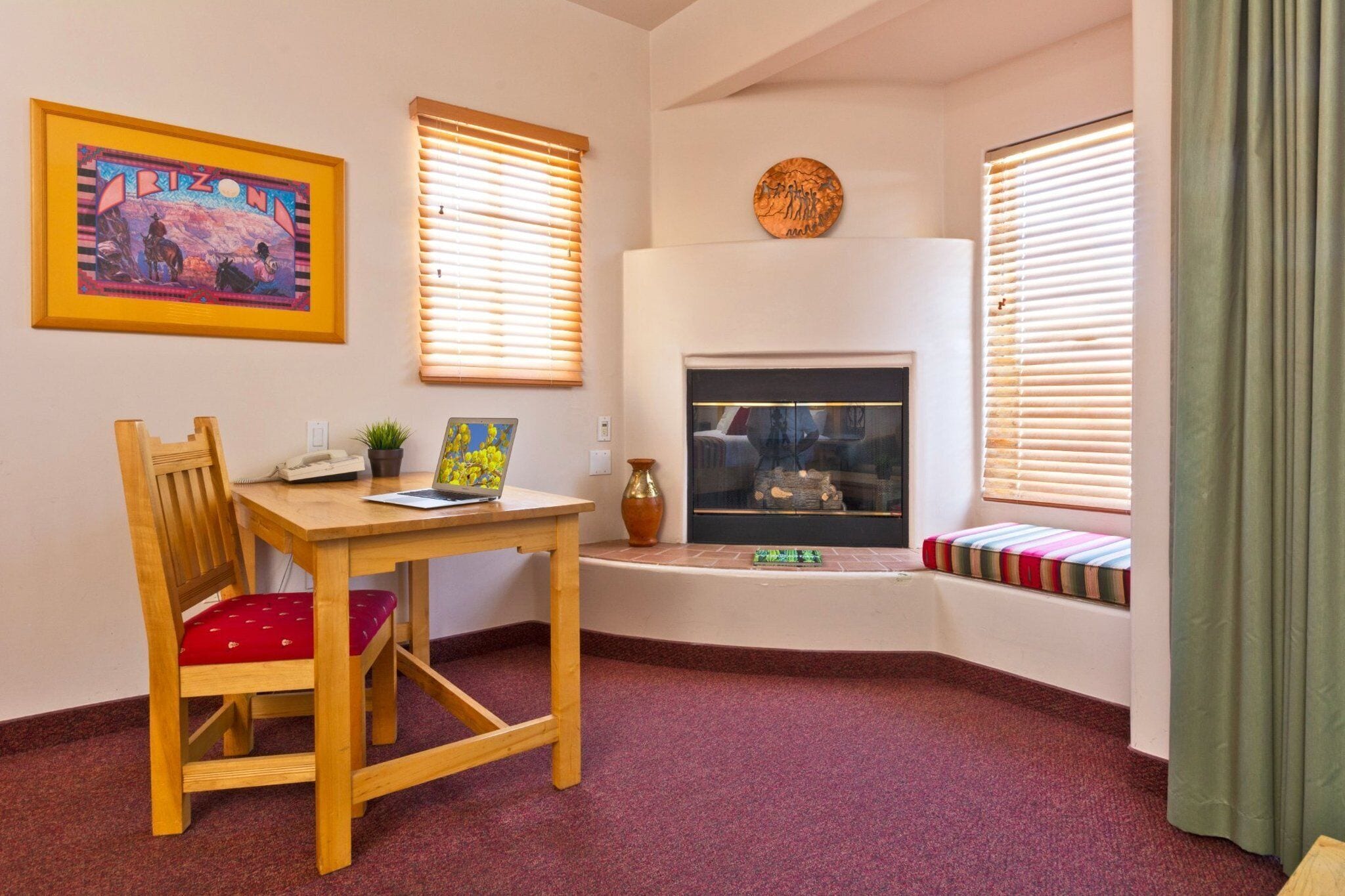 A cozy living room with a fireplace, a wooden table with a laptop, a red cushioned chair, and a window seat with a colorful cushion.