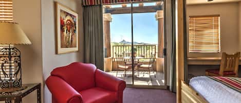 A room with a red chair, a bed, a table with a lamp, a painting, and a balcony with a view of the mountains.