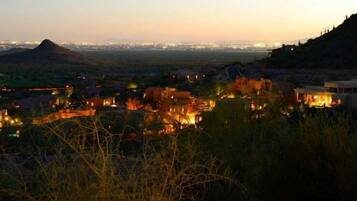 A view of a town at dusk with buildings illuminated, a hill in the background, and a distant city skyline.