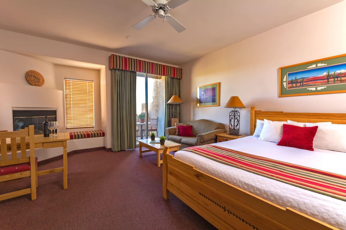 A hotel room with a bed, a desk, a chair, a sofa, a window with curtains, a ceiling fan, and a painting on the wall.