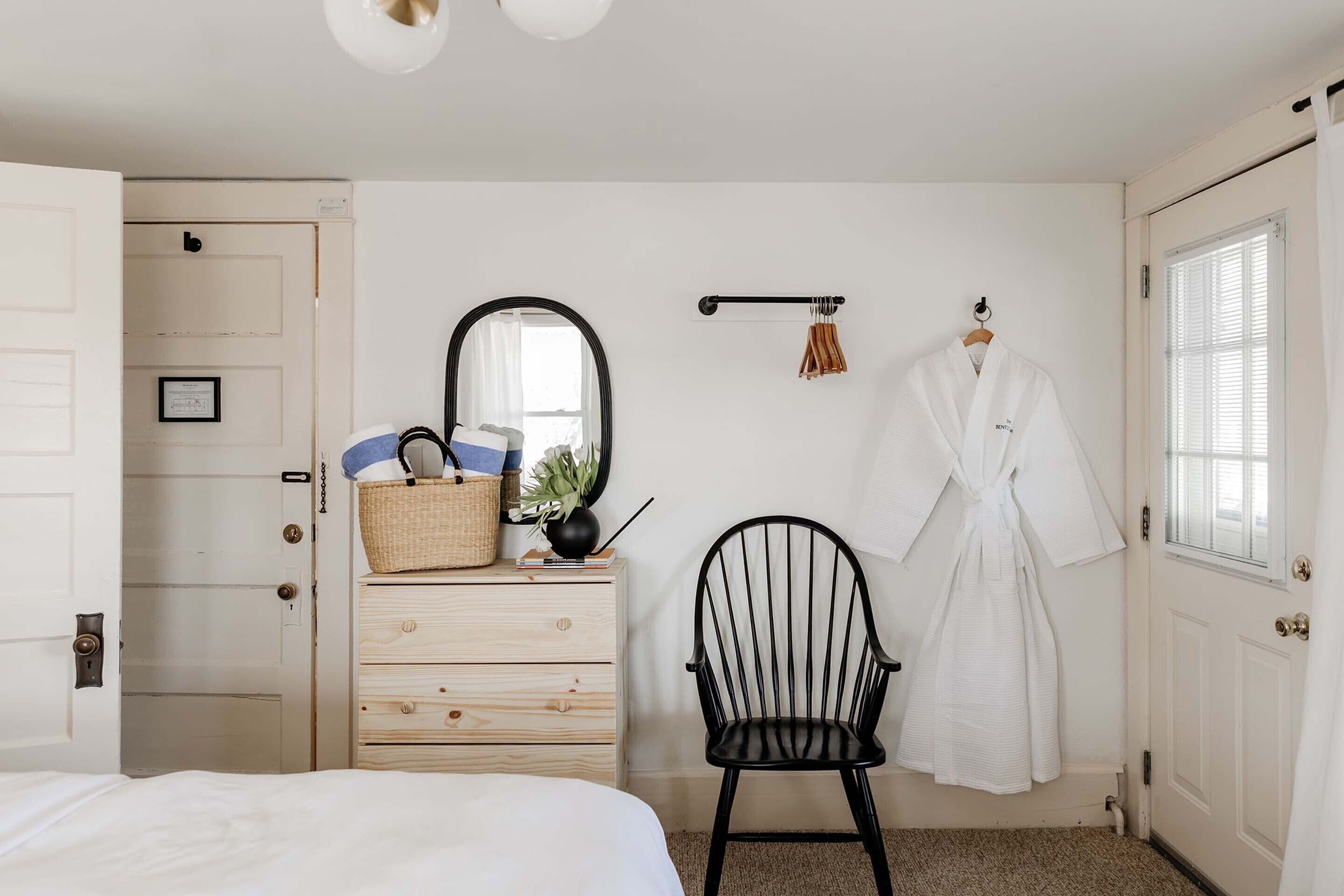 Ein Schlafzimmer mit einem Holzkasten, einem schwarzen Stuhl, einem weißen Bademantel, der an der Wand hängt, und einem Fenster mit Jalousien.