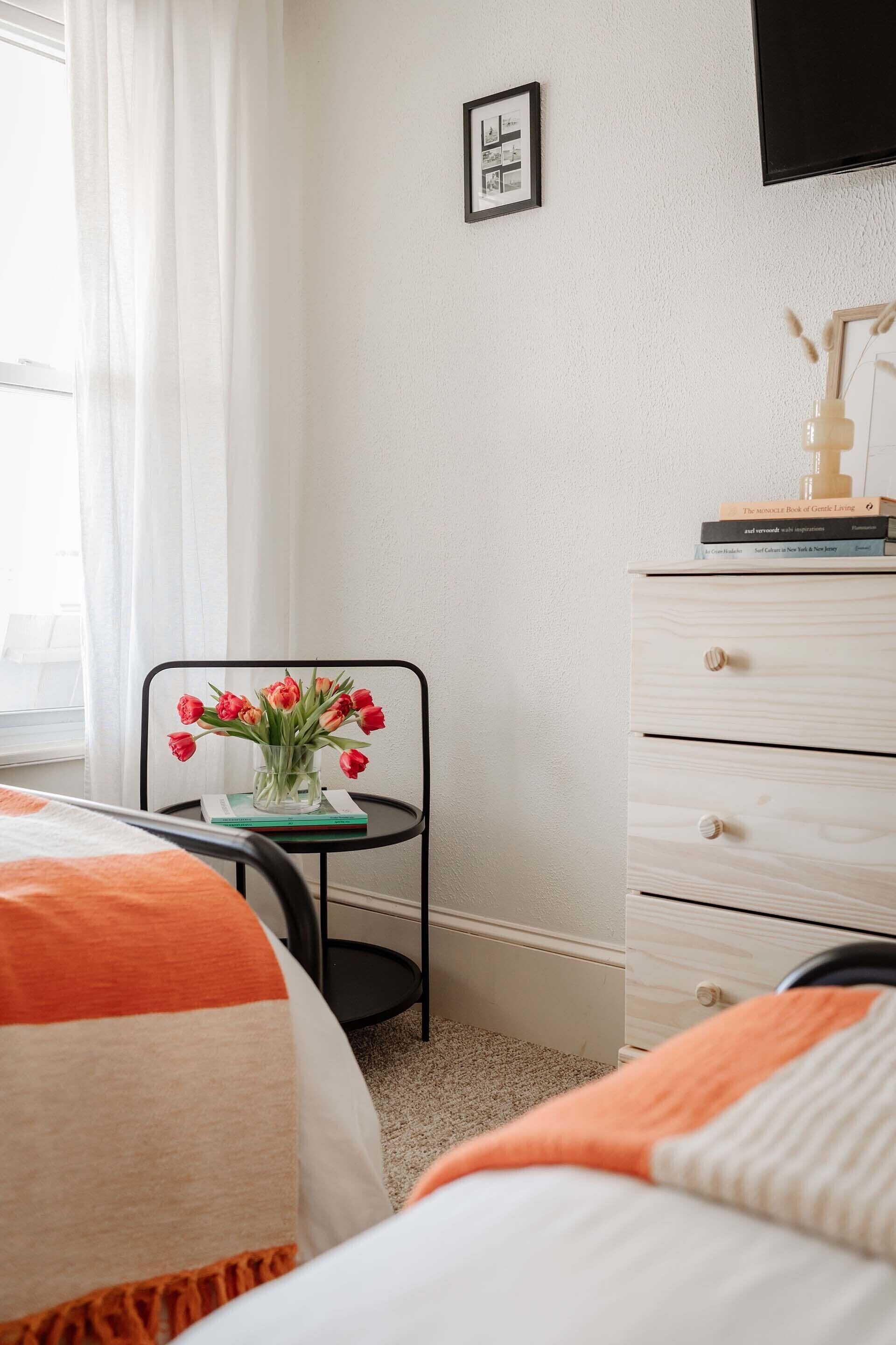 A hotel room with a bed, a small table with a vase of flowers, a dresser, and a framed picture on the wall.