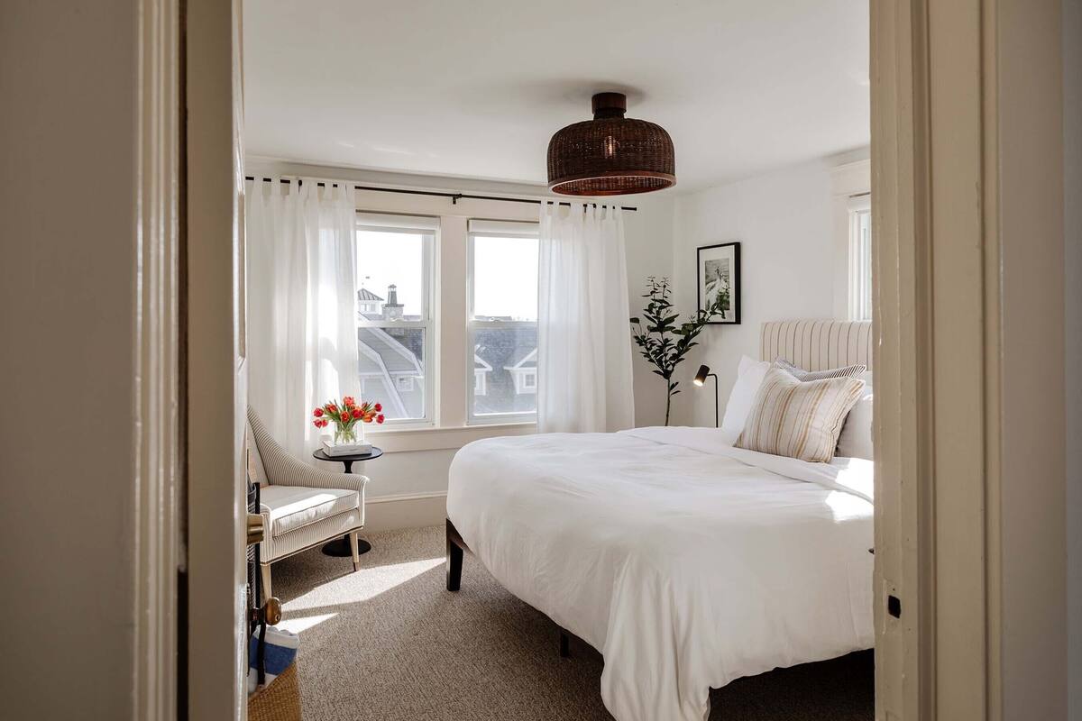 A bedroom with a large bed, a chair with a vase of flowers, and a window with curtains.