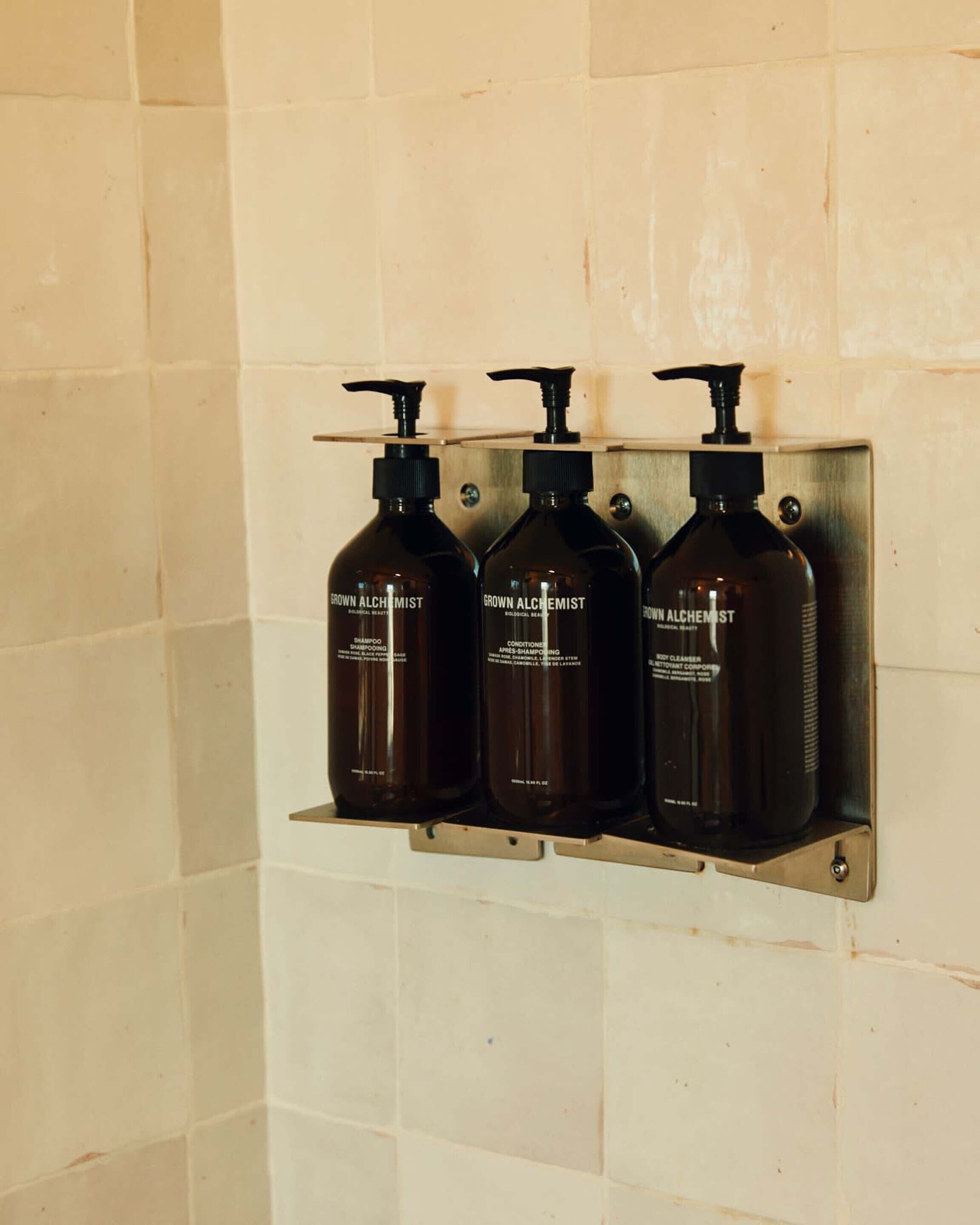 Three bottles of 'Grown Alchemist' shampoo and conditioner on a metal shelf against a tiled wall.