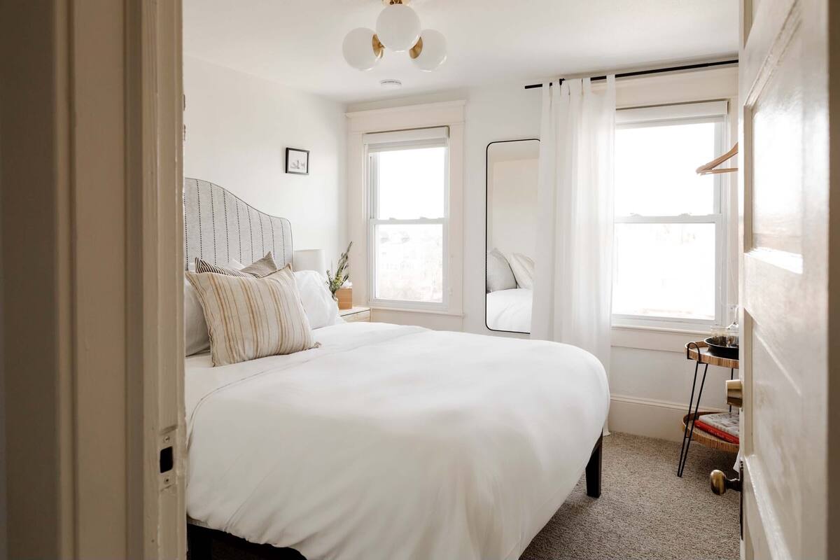 A bedroom with a large bed, a bedside table, a mirror, and a window with curtains.