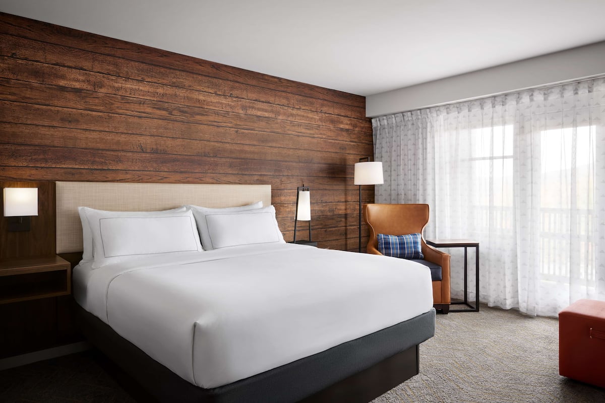 A hotel room with a large bed, wooden headboard, two bedside lamps, a chair, a small table, and a window with sheer curtains.
