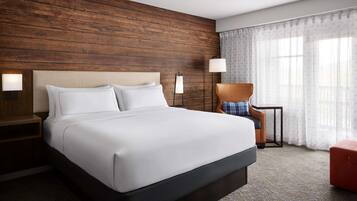 A hotel room with a large bed, wooden headboard, two bedside lamps, a chair, a small table, and a window with sheer curtains.