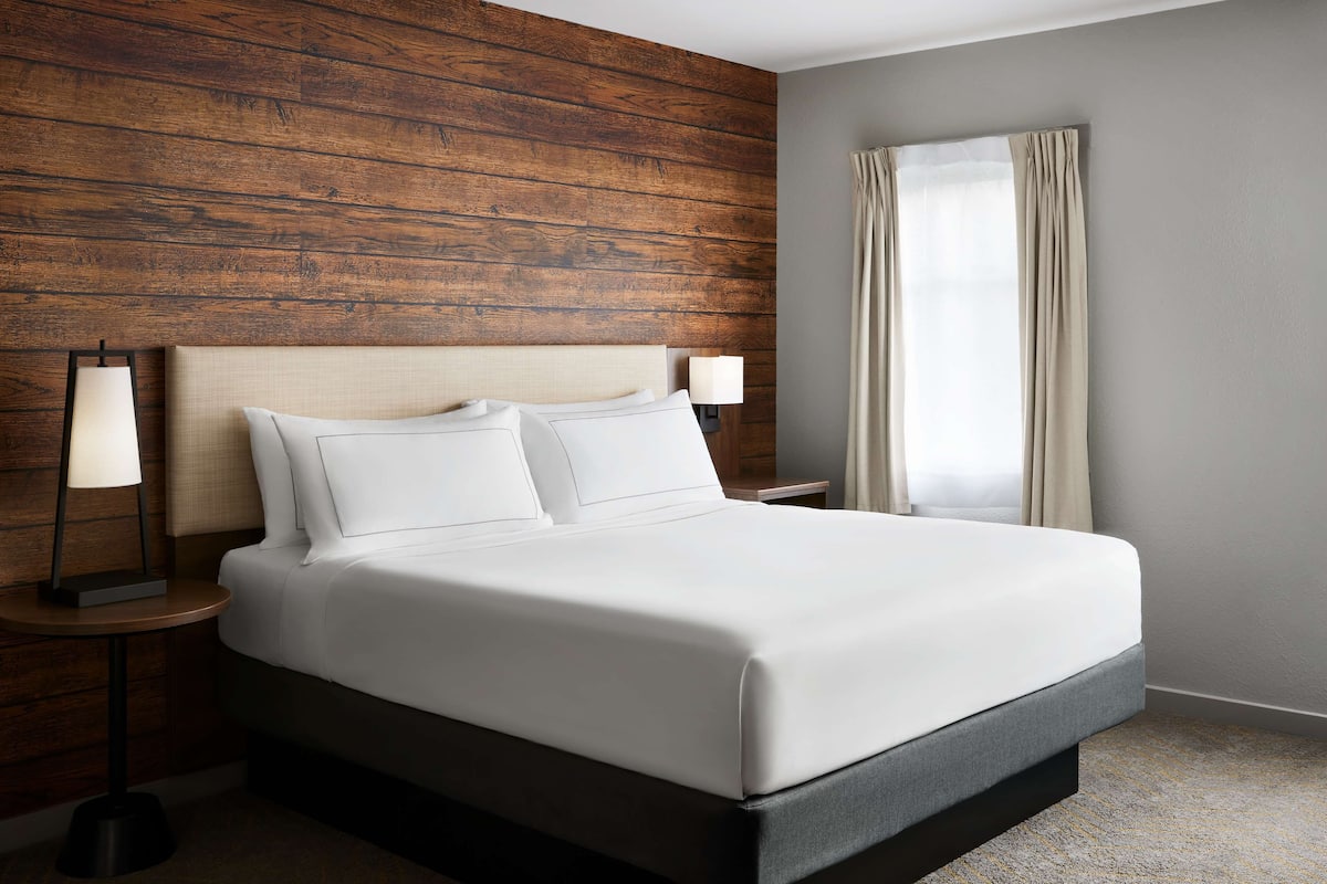 A modern hotel room with a wooden headboard, a large bed with white linens, two bedside lamps, and a window with curtains.