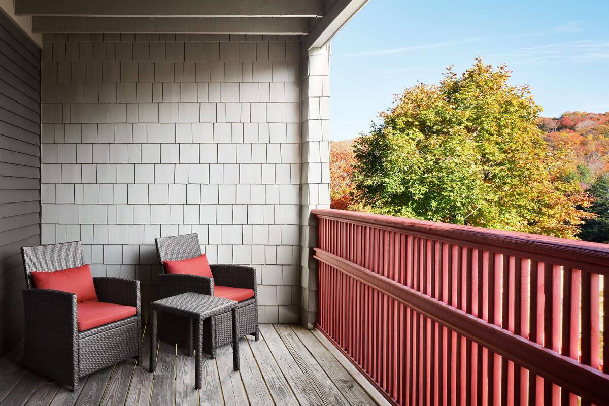A balcony with two wicker chairs and a small table, a red railing, and a view of trees with autumn foliage.
