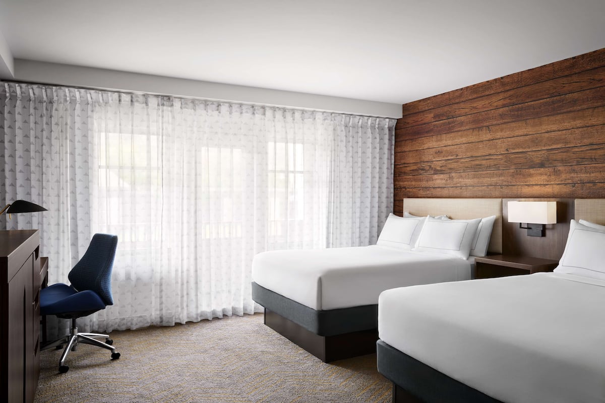 A hotel room with two beds, a desk, a chair, and a large window with sheer curtains.