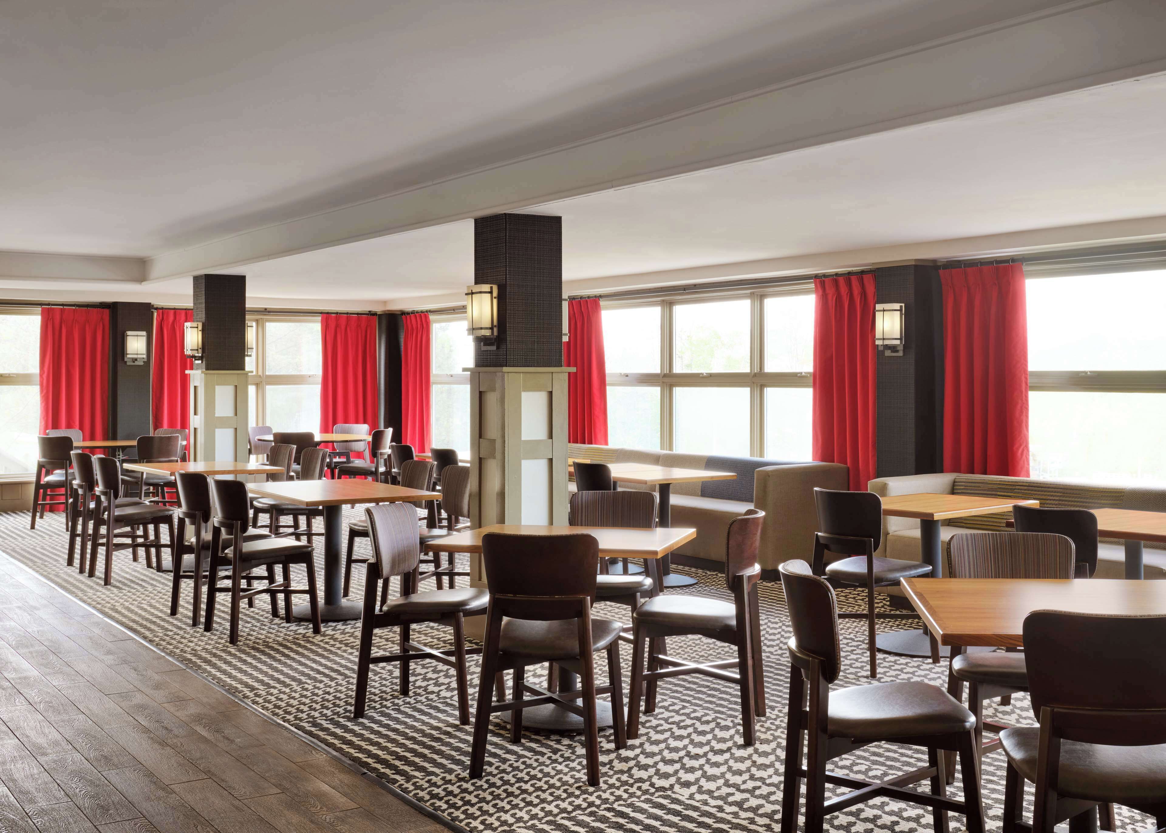 Un vaste espace dédié à la salle à manger, avec de nombreuses tables et chaises, de grandes fenêtres ornées de rideaux rouges et un pilier central.