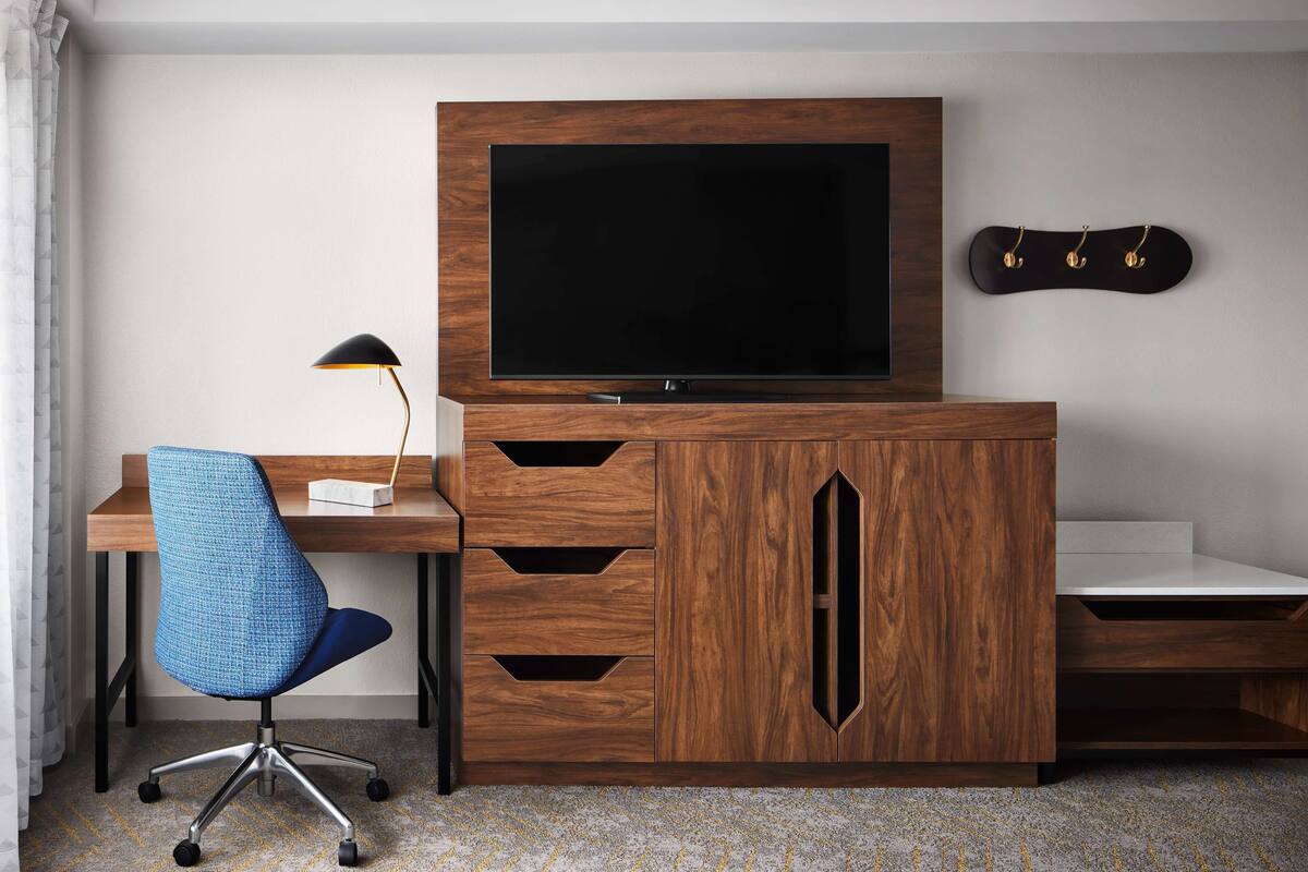 A modern hotel room with a wooden desk, a blue office chair, a flat-screen TV, and a coat rack.