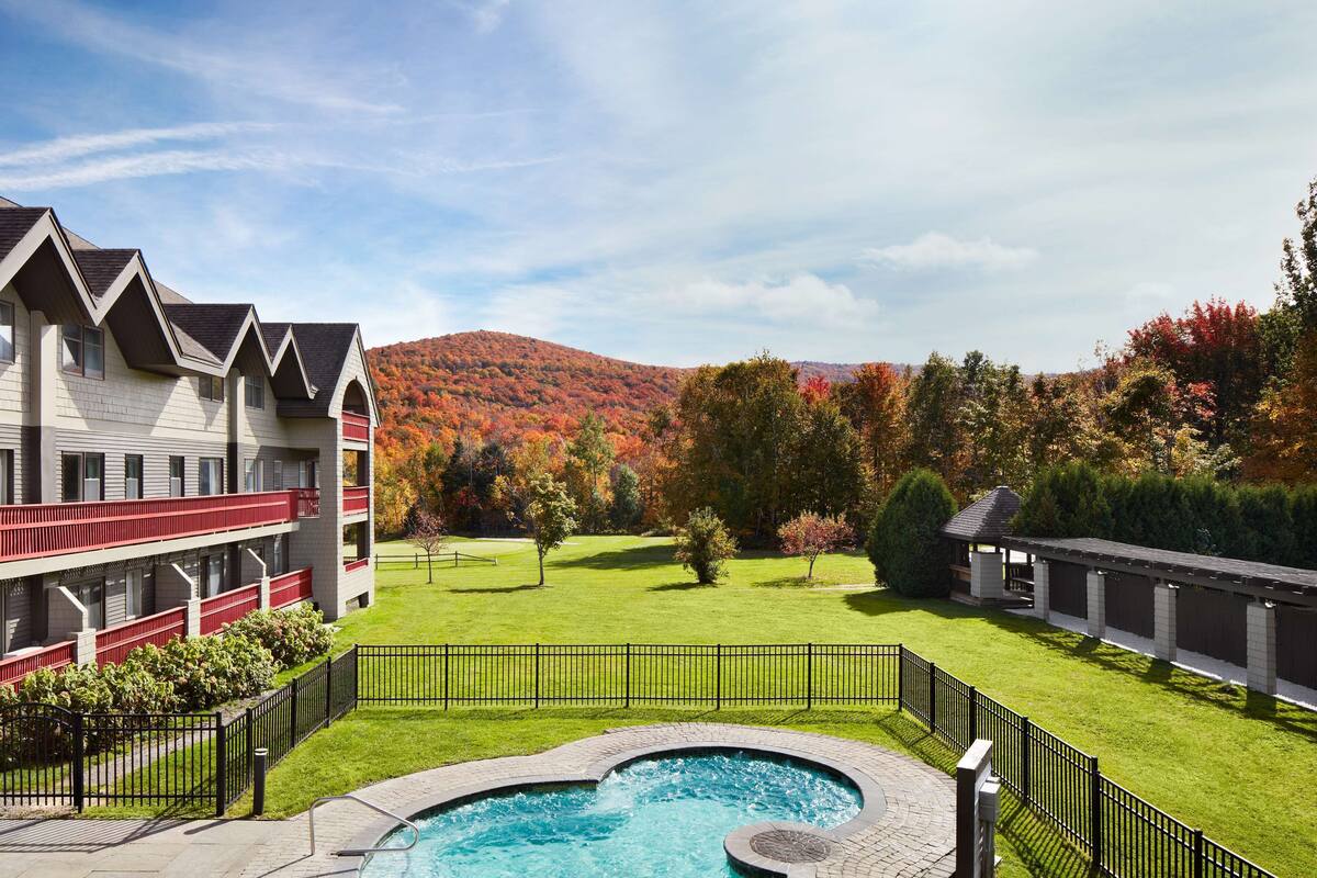 A hotel with a swimming pool, a red balcony, and a view of autumn trees and hills.