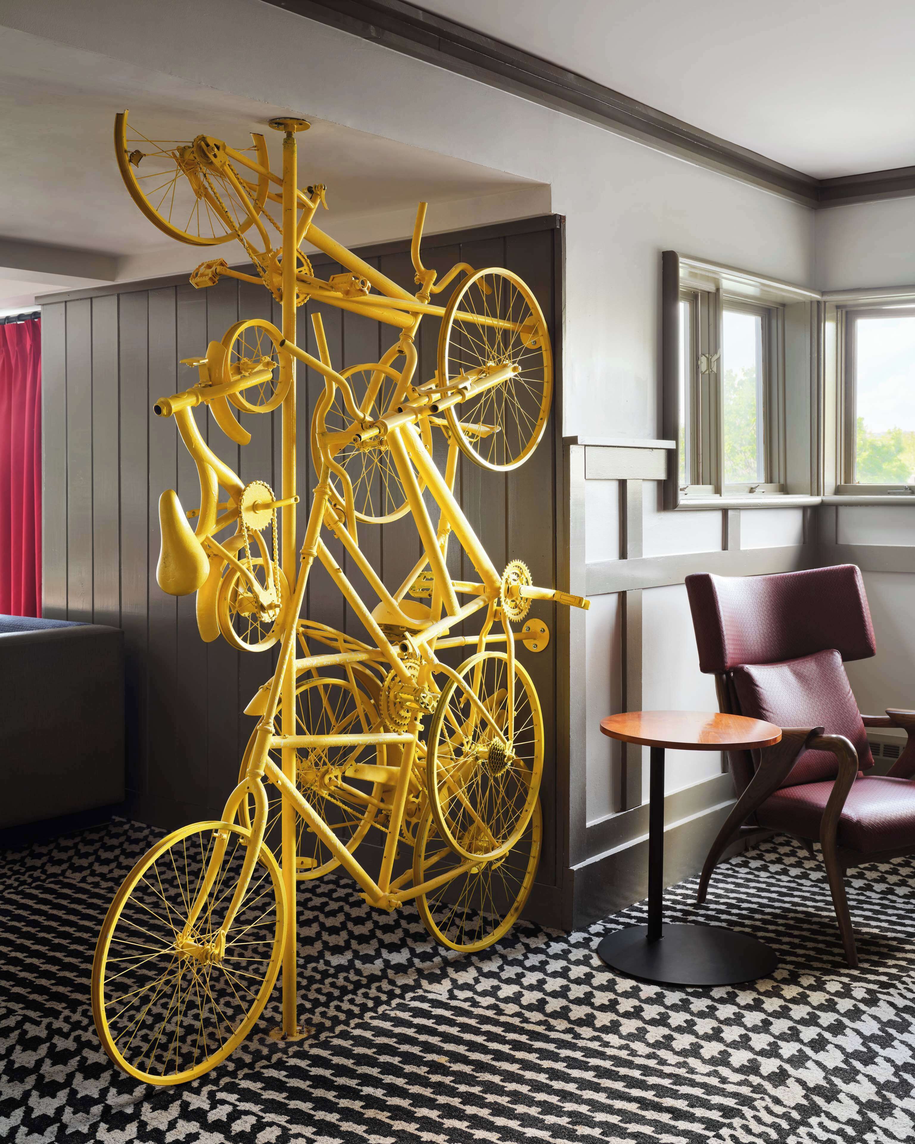 A room with a large yellow bicycle sculpture, a red chair, a patterned rug, and a window with blinds.
