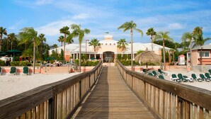 Ein hölzerner Steg führt zu einem Resort mit Liegestühlen und Palmen.