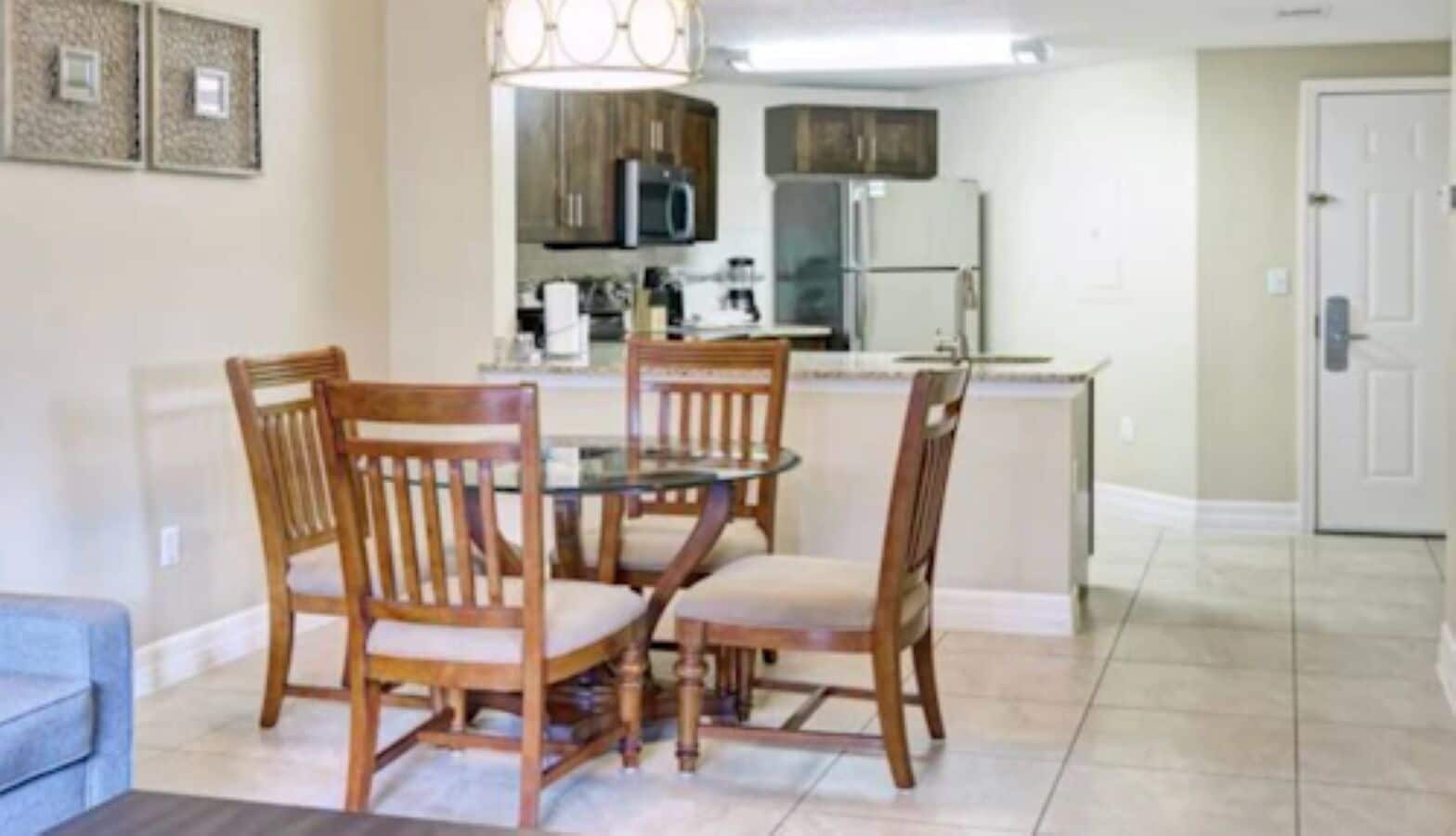 Un comedor con una mesa redonda y cuatro sillas de madera, una cocina al fondo con electrodomésticos modernos y una puerta que da a otra habitación.