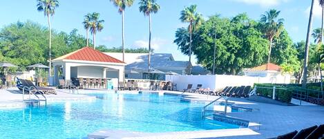 A swimming pool with lounge chairs and a covered seating area, surrounded by palm trees and a clear sky.