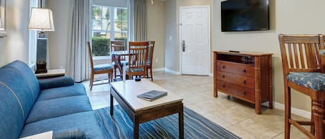 A living room with a sofa, coffee table, and chairs. There is a television on the wall and a door in the background.