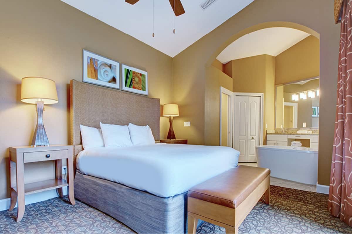 Una habitación de hotel con una cama grande, dos mesitas de noche con lámparas, un banco, un baño con espejo y una alfombra estampada.