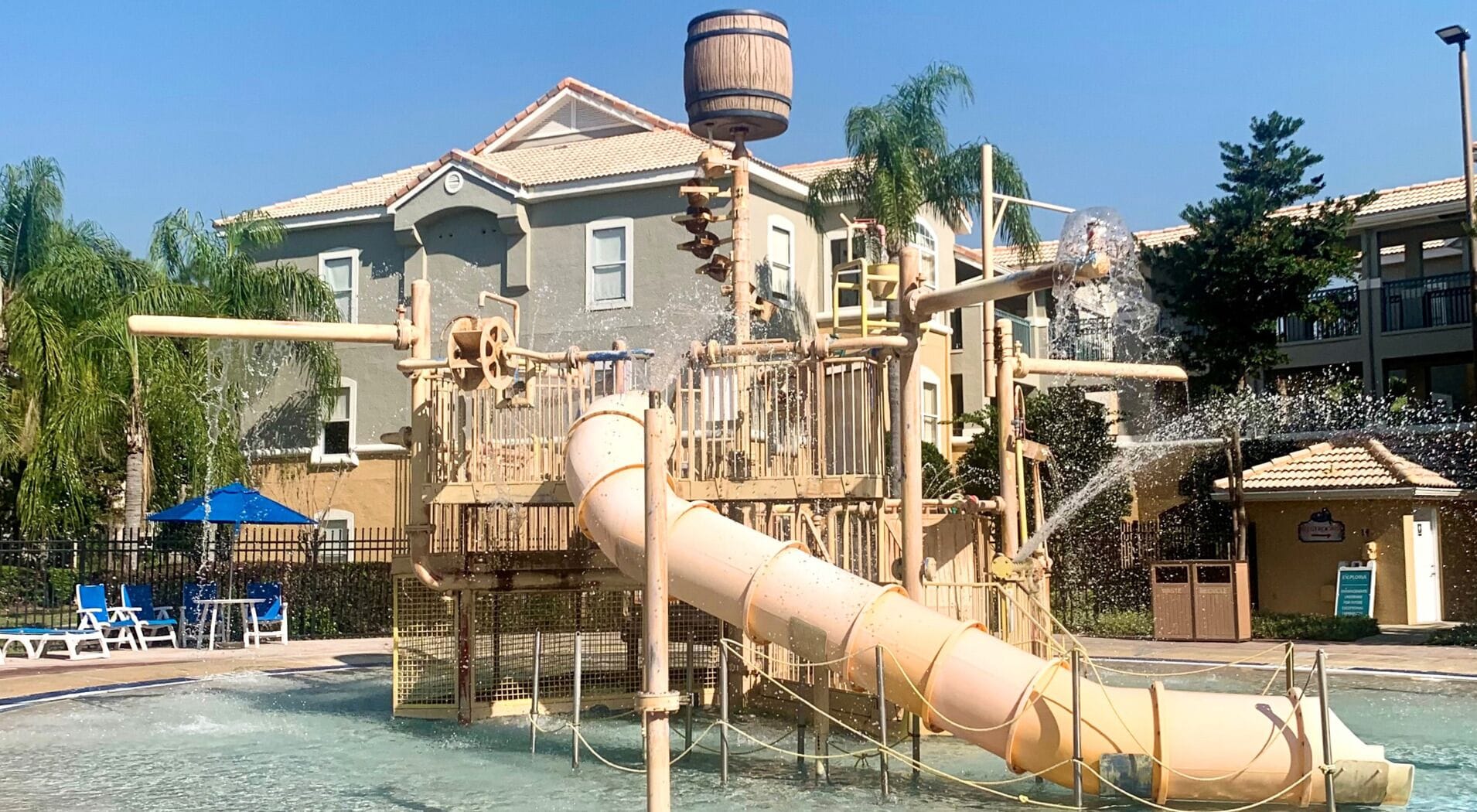 Un parque acuático con un tobogán grande, un barril de madera y una zona de piscina con sillas de descanso.