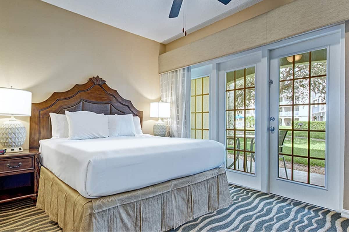 Habitación de hotel con una cama grande, dos lámparas de noche, un cabecero de madera y vistas a un área exterior por una puerta de cristal.