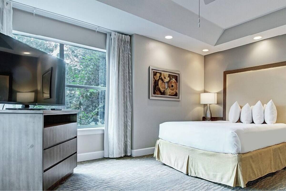 Habitación de hotel con cama, escritorio, televisión, un cuadro y ventana con cortinas.