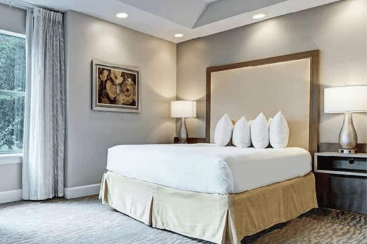Una habitación de hotel moderna con una cama grande, un cabecero, dos mesitas de noche con lámparas, una ventana con cortinas y un cuadro en la pared.
